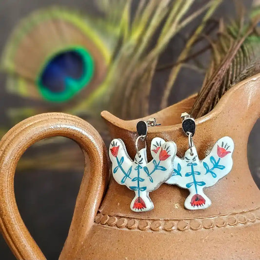 Boucles d’oreilles pendantes peintes a la main, en polymère incassables et légère, en forme d’oiseaux blanc avec motifs floraux rouges et bleus, styles folkl art, inspiration motif mexicain, présentés sur un pichet en terre cuite avec plume de paon en arrière-plan