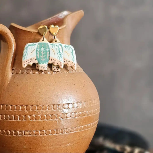 Boucles d’oreilles pendantes peintes a la main, en polymère incassables et légère, en forme d’oiseaux blanc avec motifs floraux beige et bleus, styles folkl art, inspiration motif mexicain, présentés sur un pichet en terre cuite avec plume de paon en arrière-plan