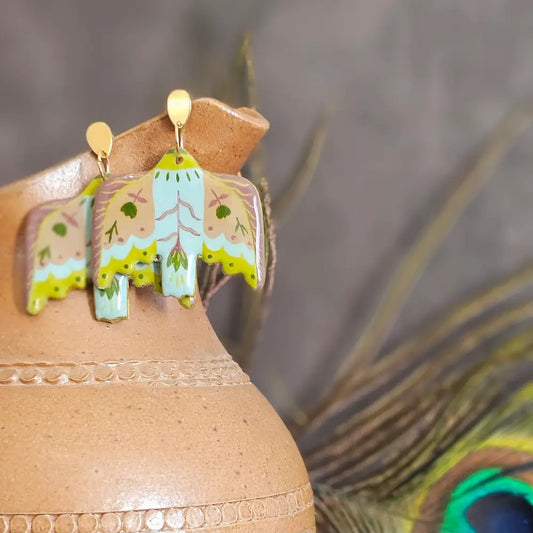Boucles d’oreilles pendantes peintes a la main, en polymère incassables et légère, en forme d’oiseaux blanc avec motifs floraux vert et brun, styles folkl art, inspiration motif mexicain, présentés sur un pichet en terre cuite avec plume de paon en arrière-plan