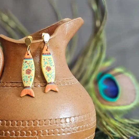 Boucles d’oreilles pendantes peintes a la main, en polymère incassables et légère, en forme de poisson jaune avec motifs floraux vert et orange, styles folkl art, inspiration motif mexicain, présentés sur un pichet en terre cuite avec plume de paon en arrière-plan