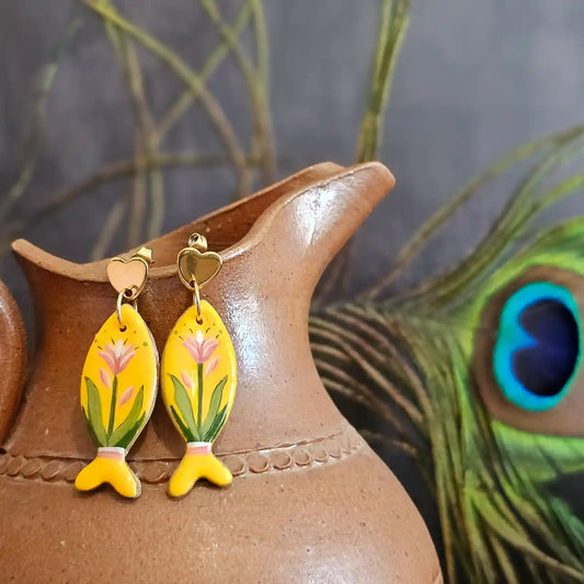 Boucles d’oreilles en forme de poisson faite a la mains. Motif floraux sur un fond jaune peint a la main. Présenté sur un pichet en argile avec une plume de paon dans le fond
