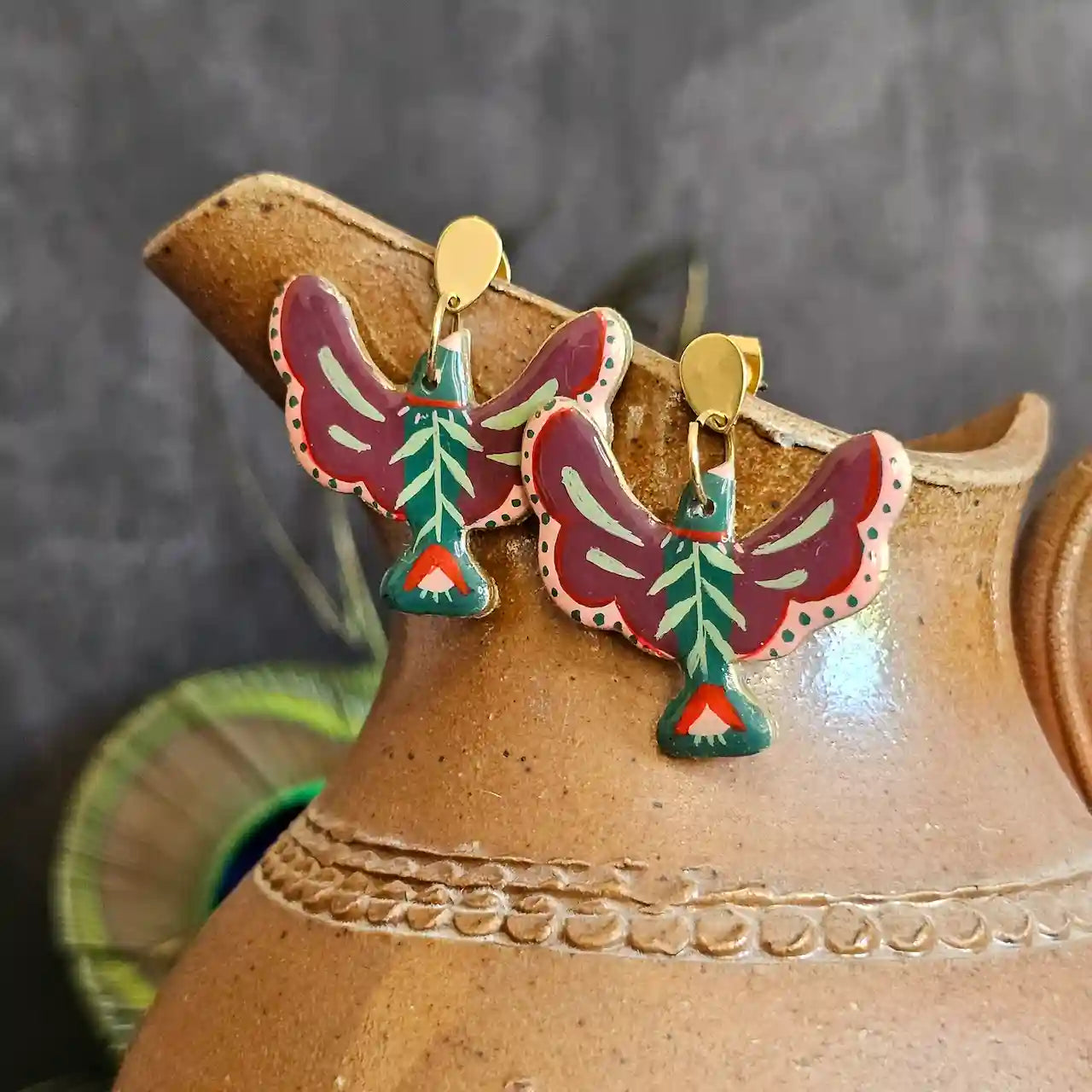 boucles d’oreilles en forme d’oiseau faites main. Oiseaux peints a la min style folk mexicain et floraux. Présenter sur un pichet en terre cuite et une plume de paon en fond