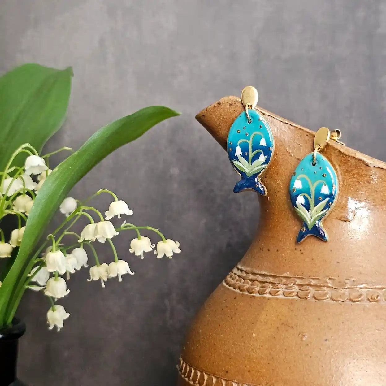 boucles d’oreilles en forme de poisson faite a la main. Motif floraux, fleur de muguets sur un fond bleues peintes a la main. Présenter sur un bichet en terre cuite avec des brun de muguets a coté 