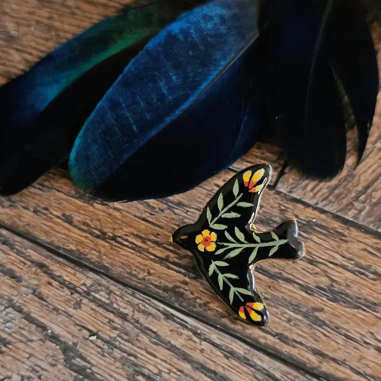 pins en foorme d’oiseau faites a la main. Motif floraux, folk mexicain, sur un fond noir. Présenter sur un fond en bois avec des plumes de paon 